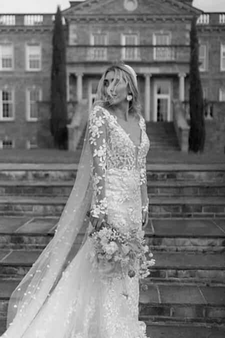 Elegant Bride In A Sheer Long-sleeved Floral Lace Wedding Gown With Flowing Cathedral Veil, Styled With Loose Romantic Waves And Statement Earrings, Holding A Wild Garden Bouquet Against Historic English Manor House Steps, Captured In Timeless Black And White.