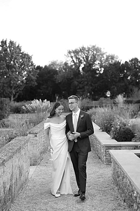 Bride Wearing Effortless Glowing Makeup With Defined Brows, Long Lashes And Sleek Centre-parted Hair, Styled In A Modern Off-shoulder Draped Wedding Dress, Photographed In A Romantic Walled Garden With Ornamental Grasses