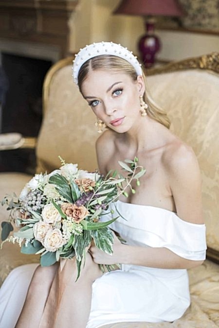 A Chic Bride With Sleek, Glowing Makeup And A Polished Low Ponytail Sits Elegantly On A Vintage Sofa, Holding A Bouquet Of Muted Peach And Ivory Flowers. She Wears A Pearl-embellished Headband And Statement Earrings, Exuding Modern Luxury With A Timeless Touch. A Perfect Inspiration For Brides Seeking A Sophisticated Yet Fashion-forward Wedding Look.