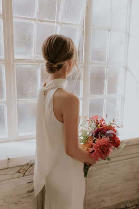 A Stylish Bride With A Sleek Low Bun Gazes Out Of A Sunlit Window, Holding A Vibrant Bouquet. Her Minimalist Yet Sophisticated Bridal Hairstyle Enhances The Contemporary Elegance Of Her Halter-neck Gown. A Perfect Look For Fashion-forward Brides Wanting Timeless Beauty With A Modern Edge.