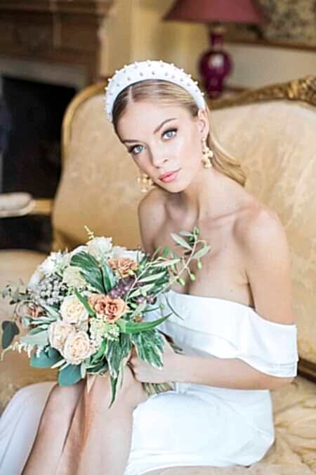 A Chic Bride With Sleek, Glowing Makeup And A Polished Low Ponytail Sits Elegantly On A Vintage Sofa, Holding A Bouquet Of Muted Peach And Ivory Flowers. She Wears A Pearl-embellished Headband And Statement Earrings, Exuding Modern Luxury With A Timeless Touch. A Perfect Inspiration For Brides Seeking A Sophisticated Yet Fashion-forward Wedding Look.