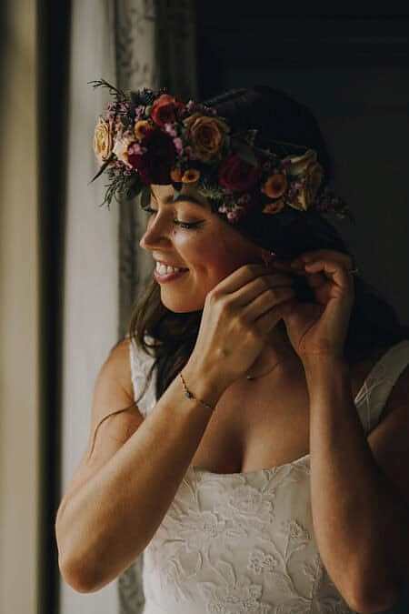 A Radiant Bride Wearing A Lush Floral Crown Smiles As She Fastens Her Earring Near A Window. Her Soft, Glowing Makeup And Natural Waves Complement Her Bohemian Bridal Style, Creating A Romantic And Effortless Wedding Look. Ideal Inspiration For Brides Seeking A Relaxed Yet Polished Aesthetic.