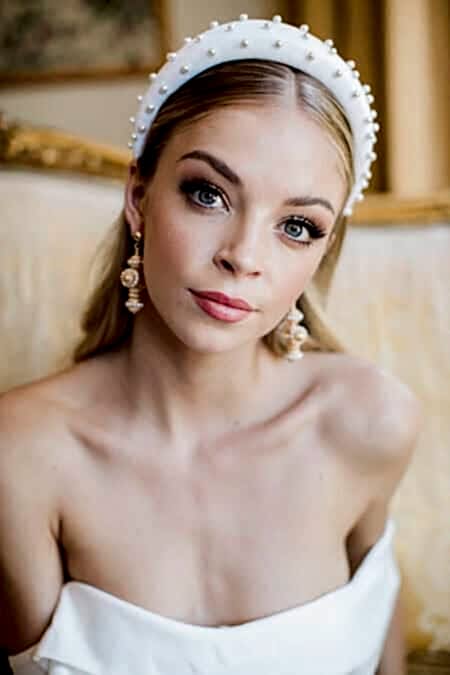 Close-up Bridal Beauty Portrait Featuring A Modern White Pearl-studded Padded Headband, Natural Glowing Makeup With Defined Lashes, Coral-pink Lips And Statement Pearl Drop Earrings, Photographed In An Elegant Period Setting With Gold-framed Mirrors.